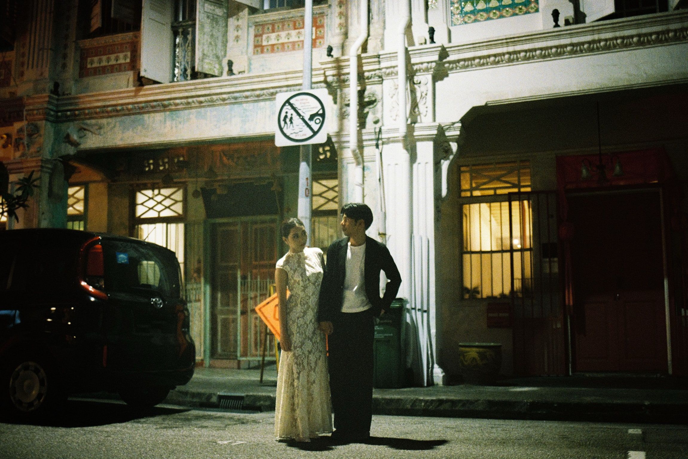 Shafiq and Syahira standing together on a shophouse street at night, warm light spilling from the doorways behind them.