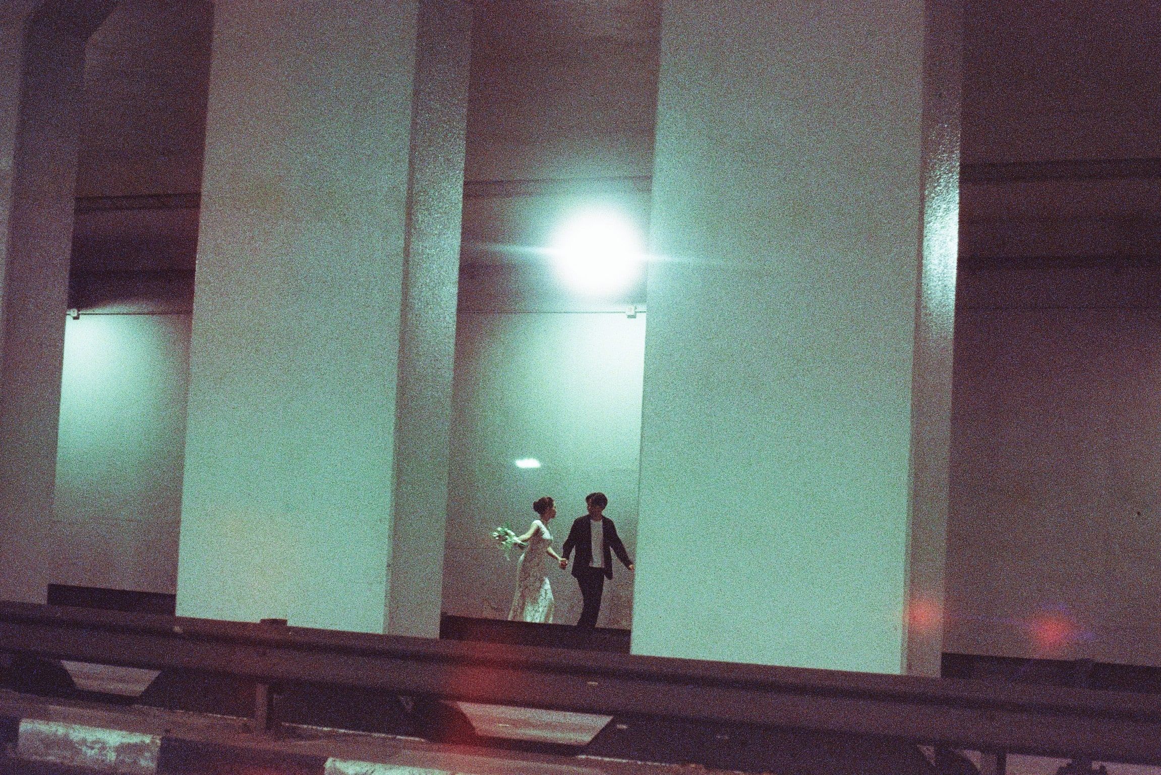 Shafiq and Syahira walking hand in hand along the lit deck of a concrete overpass at night, small between its columns.