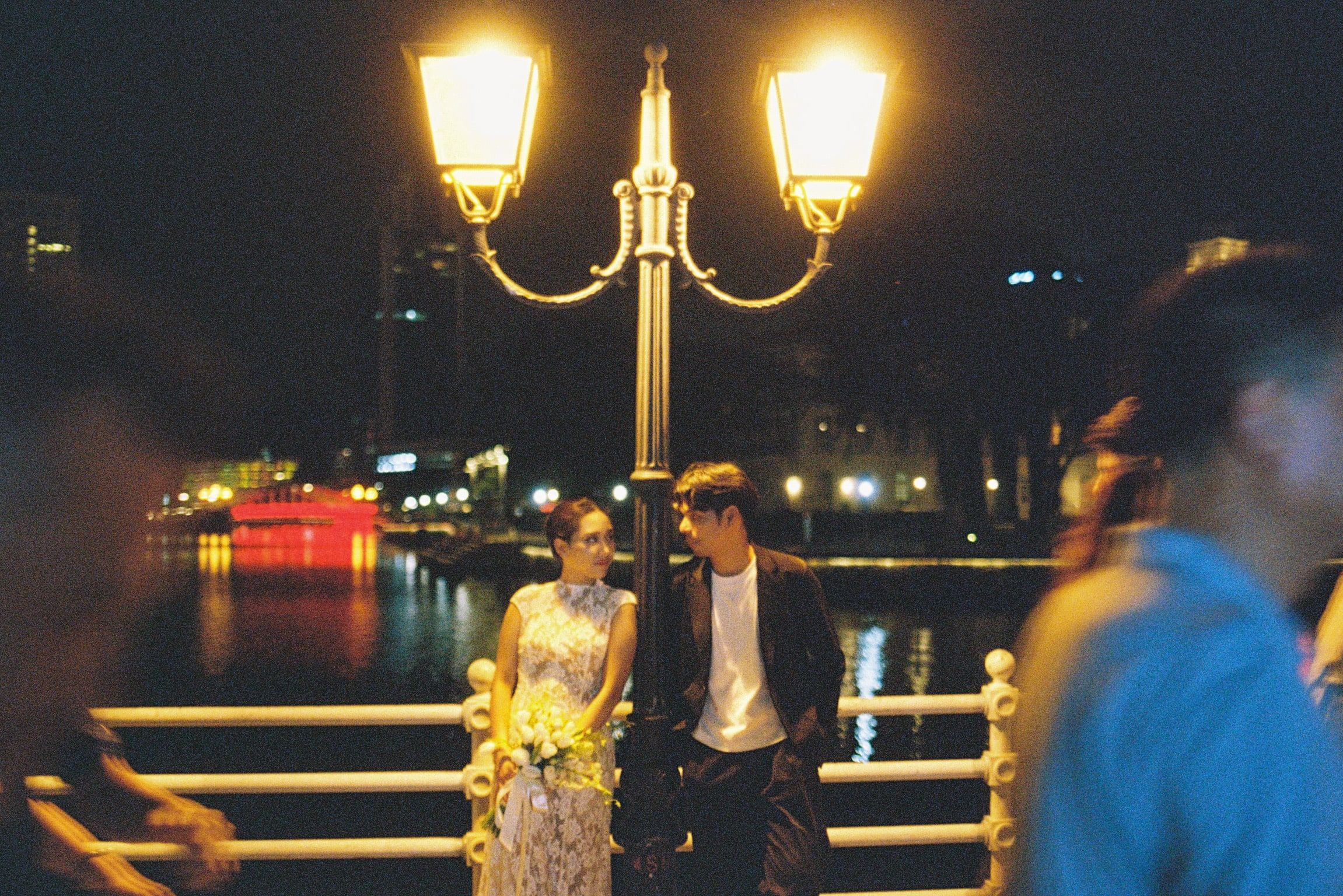 Shafiq and Syahira at a riverside railing at night beneath twin gold lamp globes, a passer-by blurred in the foreground.